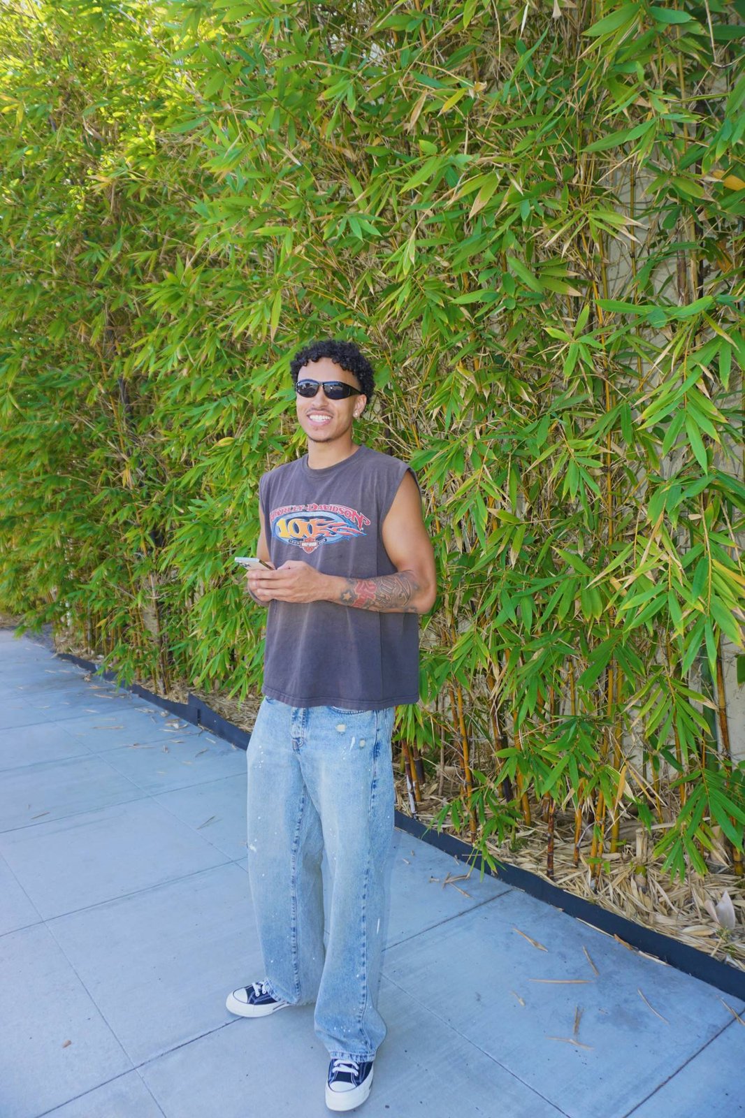 Jayo smiling in sunglasses, a vintage Harley-Davidson tank and wide-leg jeans in front of bamboo